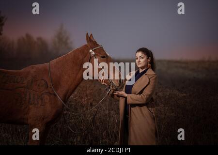 Junge Brünette Frau in einem beigen Mantel posiert mit einem rotbraunen Pferd. Sonnenuntergang Himmel und Herbst Natur. Die Inschrift auf dem Pferd: Viel Spaß. Stockfoto