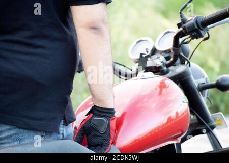 Junger indischer Mann trägt rote Reithandschuhe mit Schutz während Halten Sie die Griffstangen eines Motorrads mit einem roten Tank Stockfoto