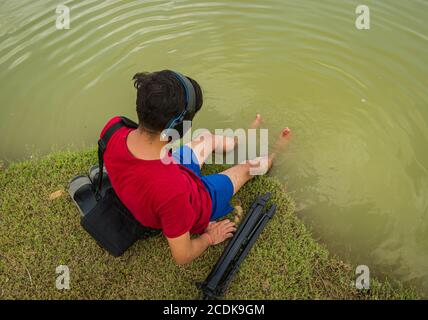 indischer Jugendlicher, der am Ufer des Sees sitzt, legt seine Füße ins Wasser, chillt, hört die Musik mit Kopfhörern und hält Handy Stockfoto