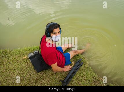 indischer Jugendlicher, der am Ufer des Sees sitzt, legt seine Füße ins Wasser, chillt, hört die Musik mit Kopfhörern und hält Handy Stockfoto