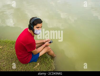 indischer Jugendlicher, der am Ufer des Sees sitzt, legt seine Füße ins Wasser, chillt, hört die Musik mit Kopfhörern und hält Handy Stockfoto