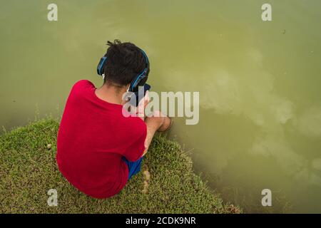 indischer Jugendlicher, der am Ufer des Sees sitzt, legt seine Füße ins Wasser, chillt, hört die Musik mit Kopfhörern und hält Handy Stockfoto