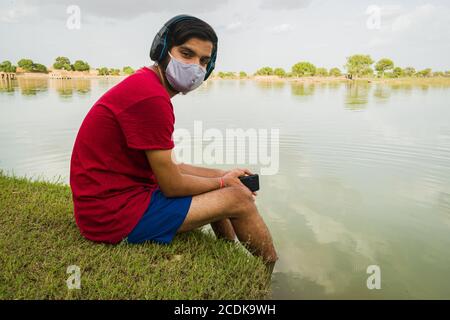 indischer Jugendlicher, der am Ufer des Sees sitzt, legt seine Füße ins Wasser, chillt, hört die Musik mit Kopfhörern und hält Handy Stockfoto