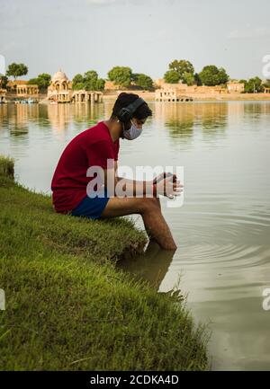 indischer Jugendlicher, der am Ufer des Sees sitzt, legt seine Füße ins Wasser, chillt, hört die Musik mit Kopfhörern und hält Handy Stockfoto