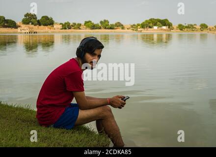 indischer Jugendlicher, der am Ufer des Sees sitzt, legt seine Füße ins Wasser, chillt, hört die Musik mit Kopfhörern und hält Handy Stockfoto