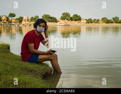 indischer Jugendlicher, der am Ufer des Sees sitzt, legt seine Füße ins Wasser, chillt, hört die Musik mit Kopfhörern und hält Handy Stockfoto