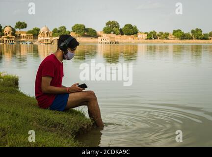 indischer Jugendlicher, der am Ufer des Sees sitzt, legt seine Füße ins Wasser, chillt, hört die Musik mit Kopfhörern und hält Handy Stockfoto