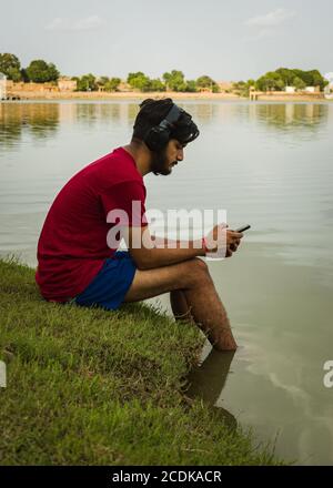 indischer Jugendlicher, der am Ufer des Sees sitzt, legt seine Füße ins Wasser, chillt, hört die Musik mit Kopfhörern und hält Handy Stockfoto