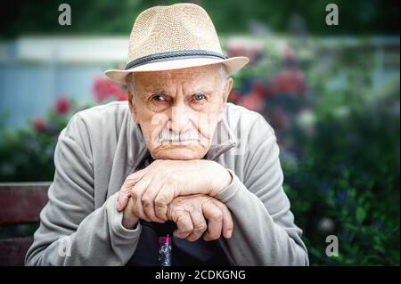 Trauriger, wütender alter Mann mit Hut sitzt in einem Freiluftgarten. Konzept von Einsamkeit und einsamem Alter Stockfoto