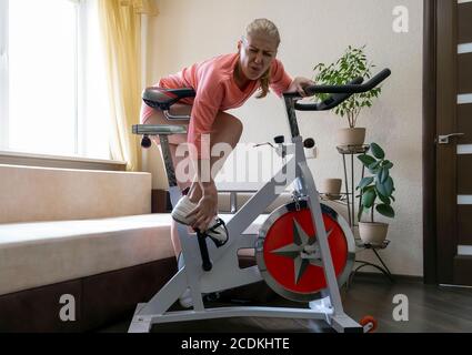 Gemeinsame Probleme. Junge Erwachsene athletische Frau verletzt, während sie ein Heimtrainer, hält sie ihr Bein und writes in Schmerzen vor dem Hintergrund eines Stockfoto