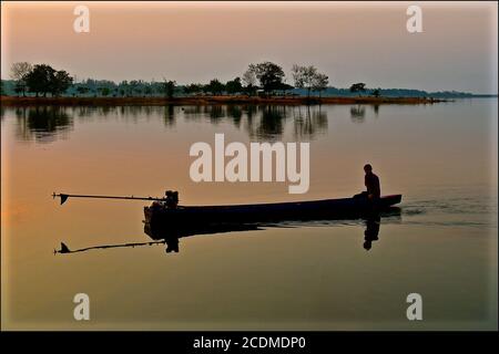 Ein Silhouettenbild eines traditionellen asiatischen Fischers in der Morgendämmerung In einem kleinen hölzernen Dugout Boot auf einer ruhigen ruhigen see mit noch Schatten Reflexionen/ Stockfoto