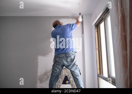 Echter Seniorenhausmaler, der auf einer Leiter steht und eine graue Wand malt. Echt dreckiger Arbeiter. Haus Maler Beruf Konzept. Stockfoto