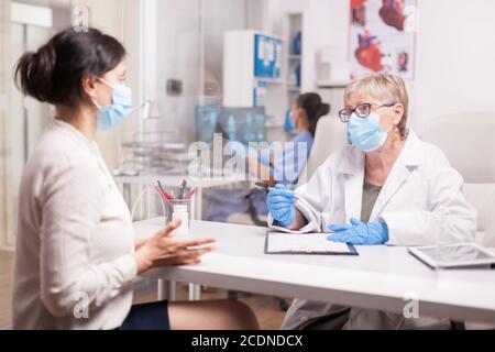 Reifer Arzt mit Gesichtsmaske gegen Coronavirus während der Konsultation mit kranken jungen Frau im Krankenhaus-Büro. Stockfoto