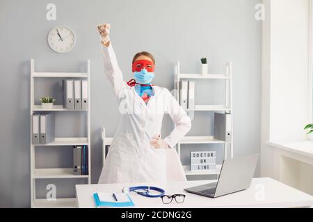 Der Arzt in einer Schutzmaske hob im Büro der Klinik die Hand in einem Superheldenanzug hoch Stockfoto