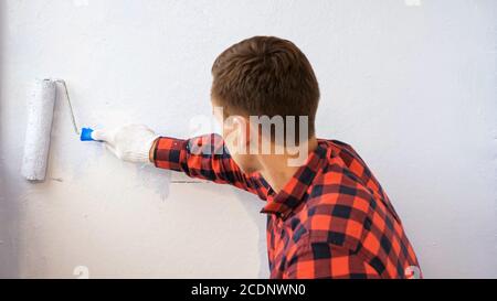 Guy in rot kariert Hemd malt Wand in hellgrau Farbe machen Reparatur auf moderne geräumige verglaste Loggia zu Hause Rückansicht Stockfoto