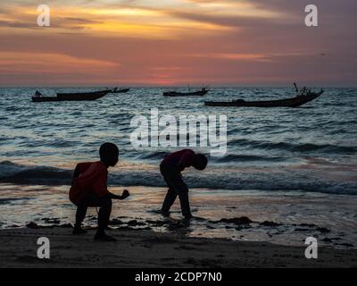 Silhouetten von Jungen, die am Strand während des Sonnenuntergangs an der Atlantikküste spielen. Im Hintergrund Silhouetten eines Fischerbooten auf dem Wasser, Senegal, Stockfoto