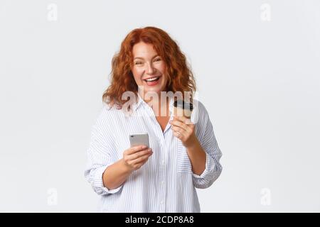 Emotionen, Lifestyle und Freizeitkonzept. Glücklich wunderschöne mittleren Alters Rotschopf Frau mit Handy und Kaffee trinken auf ihrem Weg ins Büro, suchen Stockfoto