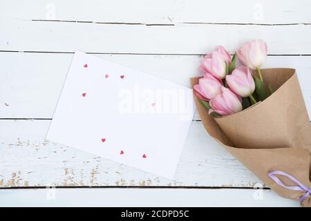 Schöner Blumenstrauß rosa Tulpen neben einem weißen Umschlag und rosa Zuckerherzen auf hölzernen Hintergrund Stockfoto