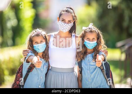 Eine Mutter und ihre Zwillingstochter benutzen eine Schutzmaske, wenn sie während der COVID-19-Quarantäne zur Schule zurückkehren und den Daumen nach oben zeigen. Stockfoto