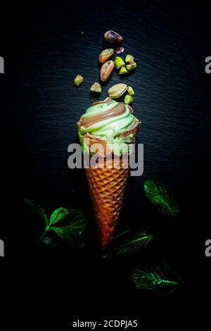 Minze Pistazien-Eis mit Schokolade in Waffelkegel, verstreute Pistazien und Minzblätter, dunkler Steinhintergrund, selektiver Fokus Stockfoto