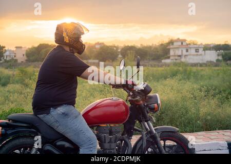 Junger inder mit Integralhelm und Schutzhandschuhen Reiten auf einem roten königlichen enfield bei Sonnenuntergang goldene Stunde Stockfoto