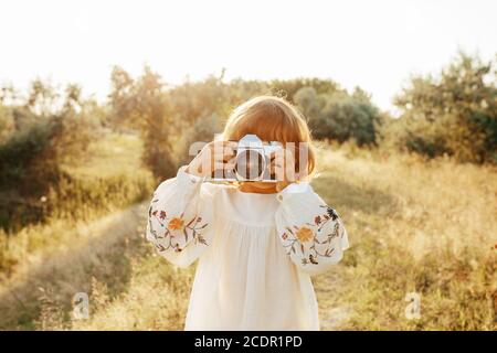 Kleines Mädchen in Stickerei Bluse, rustikalen Stil Kleid, Fotografieren mit Retro-Fotokamera hinterleuchtet Sommer Sonnenuntergang, positive Vibes, ländlichen einfachen Leben. Stockfoto