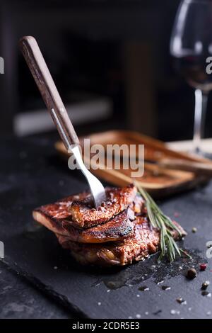 Gabel in Steak auf schwarzem Teller mit Rosmarin und Pfeffer, selektiver Fokus. Düster und launisch Stockfoto