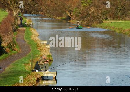 Chichester Canal von der Hunston Bridge. Mann mit Spaniel Hund, Mann Angeln, Mann Ruderboot. West Sussex, England, Dezember. Stockfoto