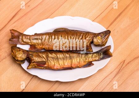 Zwei geräucherte Fische, Bachforelle, auf einem weißen Teller auf einem Buchentisch. Stockfoto