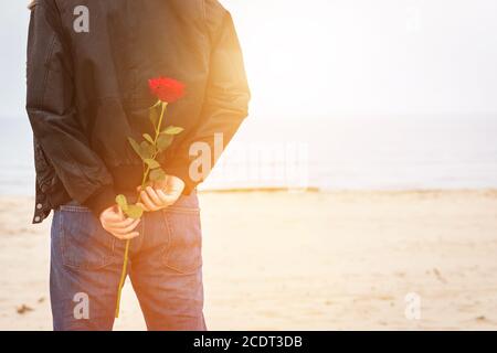 Mann mit einer Rose hinter seinem Rücken wartet auf Liebe. Romantisches Date am Strand Stockfoto