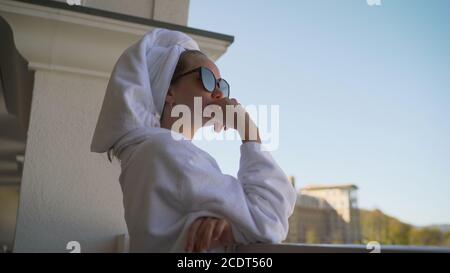 Auf dem Balkon des Hotels steht eine stilvolle Frau in einem weißen Gewand. Schöne blonde Frau in einem weißen Gewand und Handtuch, genießen die schöne Aussicht Stockfoto