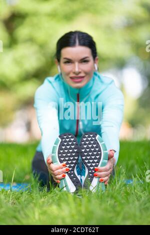 Frau erwärmt Muskeln vor dem Joggen. Sitzen auf Gras im Stadtpark - Frau dehnende Muskeln. Konzentrieren Sie sich auf die Füße. Stockfoto