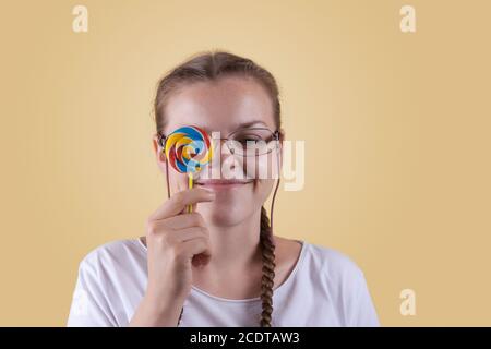 Lächelndes Mädchen hält einen Lutscher. Süßigkeiten Tag. Das Mädchen ist gut gelaunt. Farbige Lollipop für Süßigkeiten Tag. Das fröhliche Mädchen wird gegen ein gelbes b geschossen Stockfoto