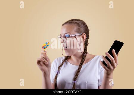 Das Mädchen schaut auf das Telefon. Das Mädchen hält das Telefon. Mädchen mit Zöpfen. Das Mädchen wird vor einem gelben Hintergrund erschossen. Das Mädchen ist überrascht b Stockfoto