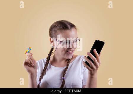 Das Mädchen schaut auf das Telefon. Das Mädchen hält das Telefon. Mädchen mit Zöpfen. Das Mädchen wird vor einem gelben Hintergrund erschossen. Das Mädchen ist überrascht b Stockfoto