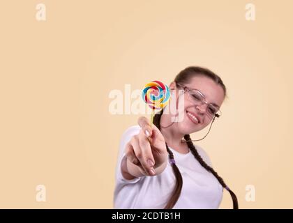 Lächelndes Mädchen hält einen Lutscher. Süßigkeiten Tag. Das Mädchen ist gut gelaunt. Farbige Lollipop für Süßigkeiten Tag. Das fröhliche Mädchen wird gegen ein gelbes b geschossen Stockfoto