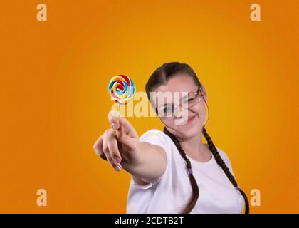 Lächelndes Mädchen hält einen Lutscher. Süßigkeiten Tag. Das Mädchen ist gut gelaunt. Farbige Lollipop für Süßigkeiten Tag. Das fröhliche Mädchen wird gegen ein gelbes b geschossen Stockfoto