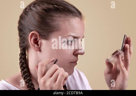 Das Mädchen schaut in den Spiegel und hält einen Pinsel. Das Mädchen trägt Make-up. Ein nettes Mädchen sieht in einem Spiegel mit einer Palette und wendet Make-up mit einem Pinsel. Stockfoto