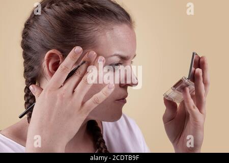 Das Mädchen schaut in den Spiegel und hält einen Pinsel. Das Mädchen trägt Make-up. Ein nettes Mädchen sieht in einem Spiegel mit einer Palette und wendet Make-up mit einem Pinsel. Stockfoto