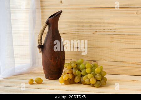 Stillleben mit grünen Trauben und einem Steingutkrug. Trauben und ein Krug auf einem hellen natürlichen Holzhintergrund. Rustikaler Stil. Warmer Vintage-Geschmack. Herbst Stockfoto