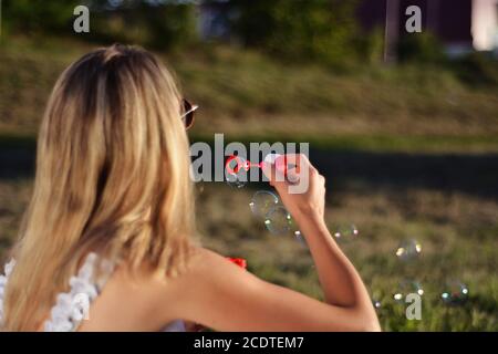 Blonde Frau bläst Seifenblasen im Freien im Park auf Sommer Sonnenuntergang. Glück in der Natur Konzept. Nahaufnahme, selektiver Fokus Stockfoto