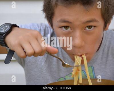 Hungriger gemischter Rasse Pre-teen Junge (Ostasiaten und Kaukasischen) isst trockene Nudeln mit einer Gabel aus einer Keramikschale. Stockfoto