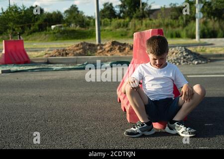 Weißer Junge sitzt auf der Straße in der Nähe des roten Zauns, weinend und sehr aufgeregt Stockfoto