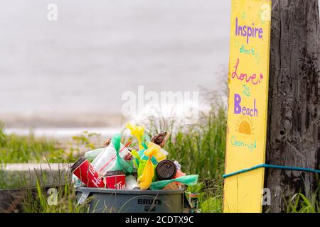 Saint John, NB, Kanada - 20. Juni 2020: Eine Mülltonne an einem Strand, der mit Müll überfüllt ist. Ein Schild ist an einen Strommast neben dem Mülleimer gebunden. Stockfoto