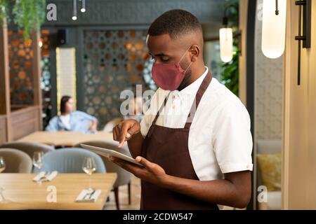 Junger afrikanischer Mann in brauner Schürze und Schutzmaske nehmen Online-Bestellungen Stockfoto