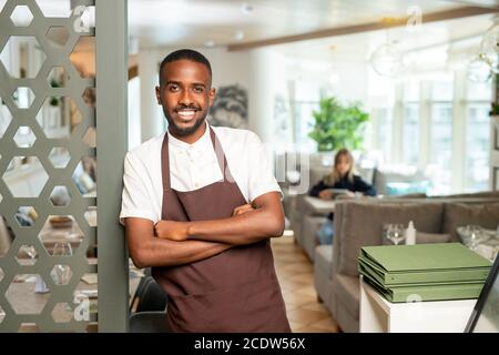 Fröhlicher junger afrikanischer Kellner in Arbeitskleidung, der sich gegen die Tür lehnte Restaurant Stockfoto