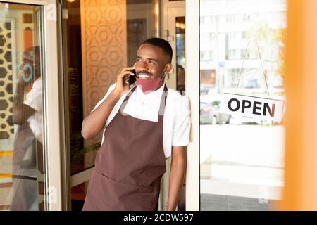 Fröhlicher junger Kellner in Uniform und Schutzmaske im Stehen Die Tür des Cafés Stockfoto
