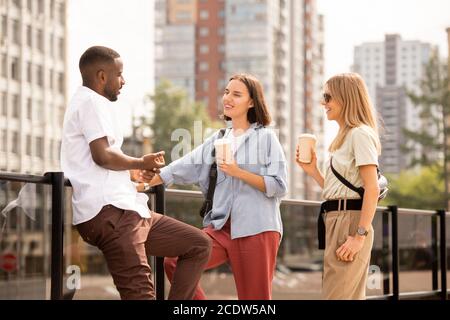 Glückliche junge blonde und brünette Frauen mit Getränken während des Gesprächs An den Afrikaner Stockfoto