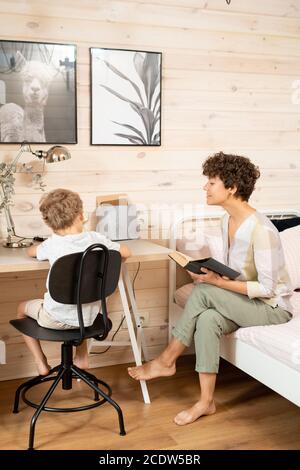 Junge hübsche Frau mit offenem Buch sitzend auf dem Bett vorbei Ihr kleiner Sohn macht Notizen Stockfoto