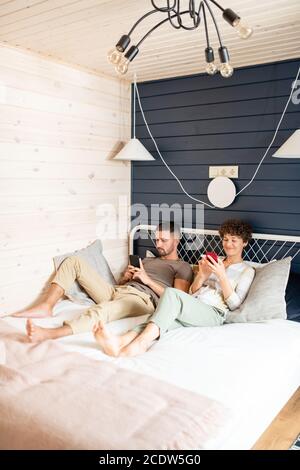 Junge glückliche Mann und Frau entspannen auf dem Bett und Scrollen In mobilen Gadgets Stockfoto